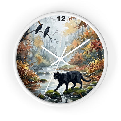 A round wall clock with a white frame and a printed autumn forest face. The artwork shows a black cat walking along a stream, two crows perched on branches, and fall foliage in yellow, orange, and brown tones.