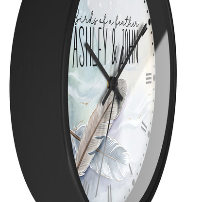 A close-up of a round wall clock with a matte black frame and a white dial featuring a feather illustration and the text 'birds of a leather ASHLEY & JOHN'.