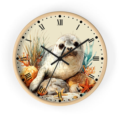 A round wall clock with a light wood frame and a whimsical illustration of a seal pup on the face, surrounded by seashells, starfish, coral, and rocks. The clock features Roman numerals and black hands on a clean white background.