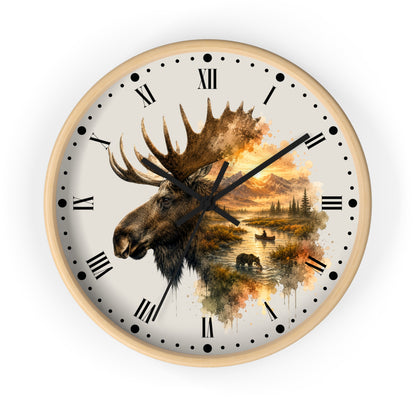 A round wall clock with a light wood frame and a cream-colored face, featuring a large moose head illustration over a scenic autumn landscape with mountains, trees, and water. The clock has black Roman numerals and black hands, shown against a plain white background.