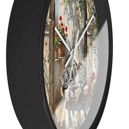 Round wall clock with a black frame and a printed scene of a gray tabby cat lounging on a sunlit stone balcony with red flowers and climbing greenery, shown against a white background.
