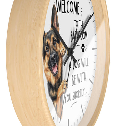A round wall clock with a light natural wood frame and a white face featuring a German Shepherd illustration and the text 'WELCOME TO THE BATHROOM A DOG WILL BE WITH YOU SHORTLY.