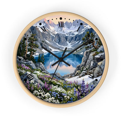 A round wall clock with a light natural wood frame and a scenic mountain landscape printed on the face. The artwork shows snow-capped peaks, a blue alpine lake, rocky cliffs, evergreen trees, and wildflowers, with black Roman numeral hour markers and black clock hands.