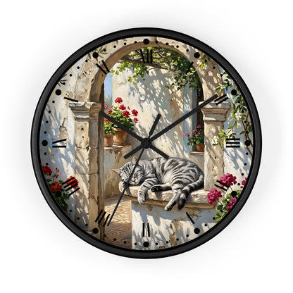 Round wall clock with a black frame and a printed scenic design featuring a gray tabby cat sleeping on a stone ledge under an arched doorway with flowers and greenery.