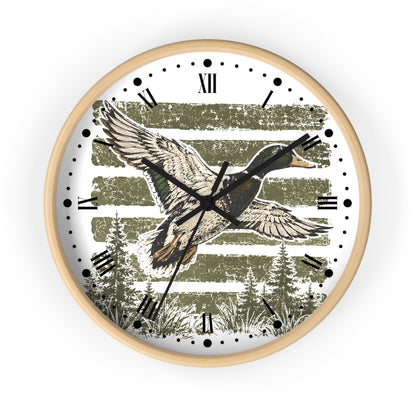 Round wall clock with a light wood frame, featuring a rustic illustration of a flying mallard duck over muted green horizontal stripes and pine trees on a white face with black Roman numerals and hands.