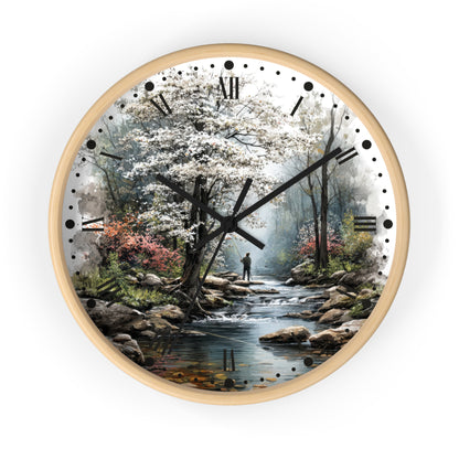 Round wall clock with a light wood frame and a scenic print of a stream, blossoming trees, rocks, and a small figure in the distance. Black Roman numerals and dot hour markers are visible on a white clock face.