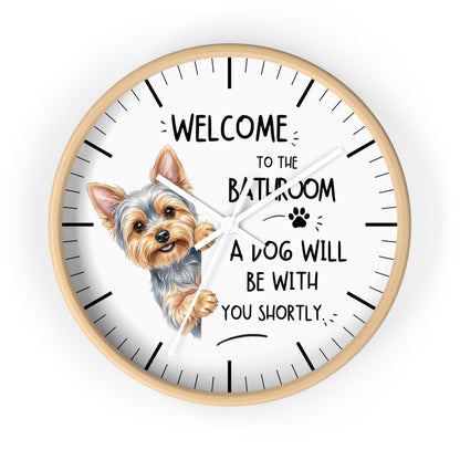 A round wall clock with a light wood frame and a white face, featuring a cartoon illustration of a Yorkshire Terrier and the humorous text 'WELCOME TO THE BATHROOM A DOG WILL BE WITH YOU SHORTLY.' on a plain white background.