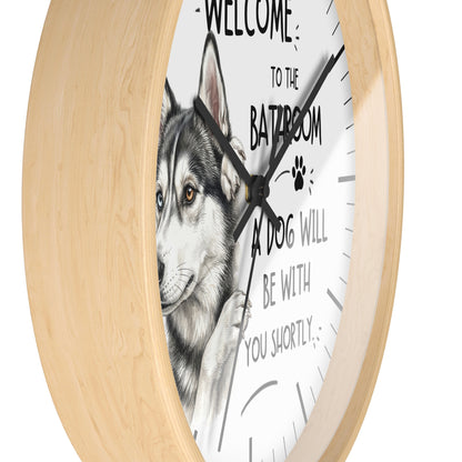 A round wall clock with a light natural wood frame and a white face featuring a husky illustration and the text 'WELCOME TO THE BATROOM A DOG WILL BE WITH YOU SHORTLY'.