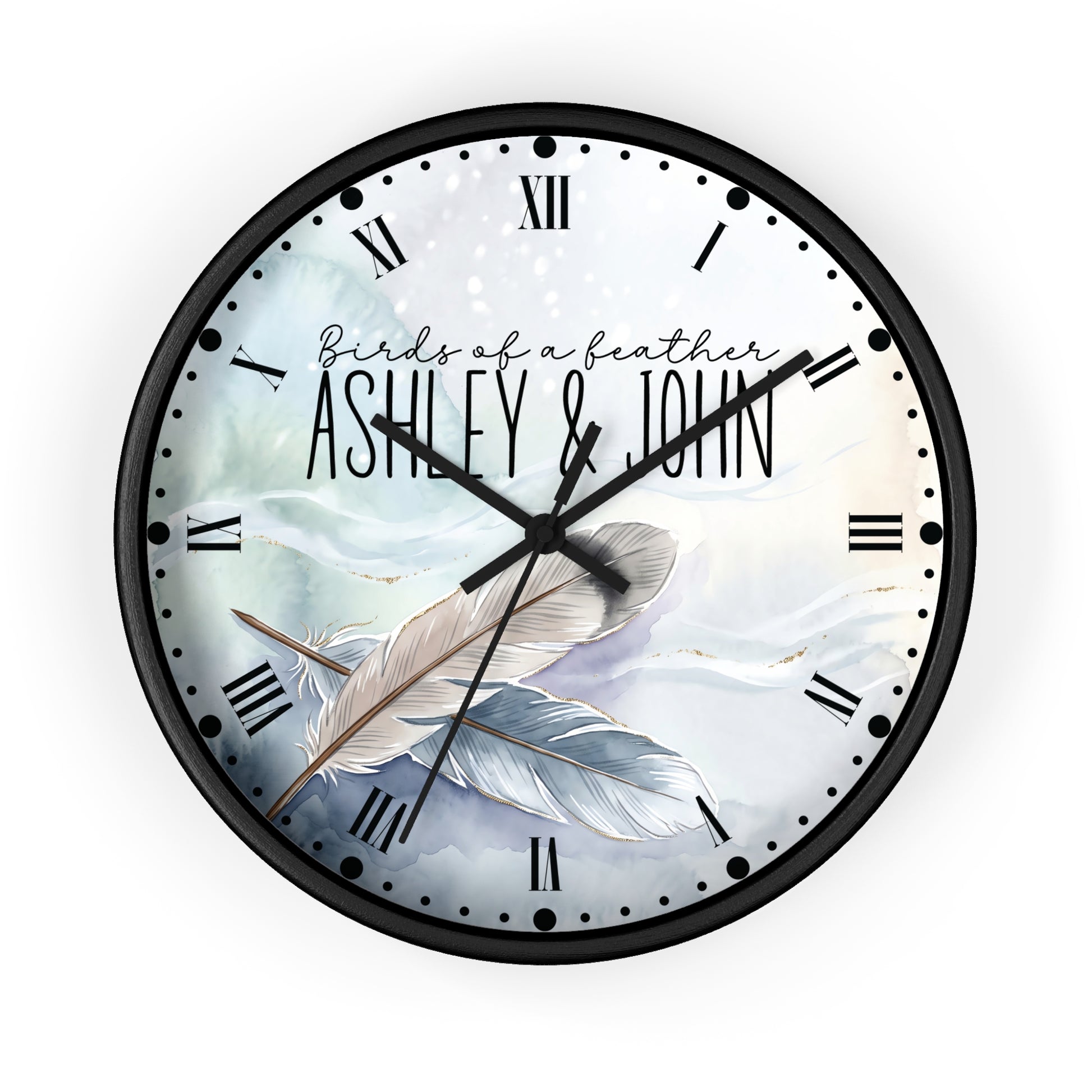 A round wall clock with a black frame and a custom printed dial featuring the text 'Birds of a feather ASHLEY & JOHN' over an illustration of two feathers on a soft blue and white background.