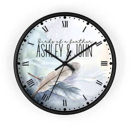 A round wall clock with a black frame and a custom printed dial featuring the text 'Birds of a feather ASHLEY & JOHN' over an illustration of two feathers on a soft blue and white background.
