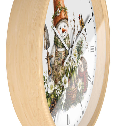 A round wall clock with a light natural wood frame and a white face featuring a whimsical winter garden illustration. The design includes a snowman wearing a gardening hat and apron, holding a trowel, with white flowers, evergreen branches, a bird, and a basket of produce.