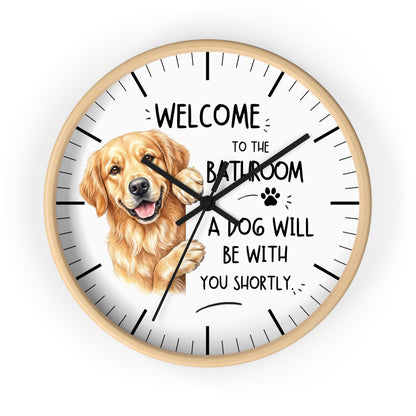 A round wall clock with a light wood frame and a white face, featuring a golden retriever illustration and the text 'WELCOME TO THE BATHROOM A DOG WILL BE WITH YOU SHORTLY.' on a plain white background.