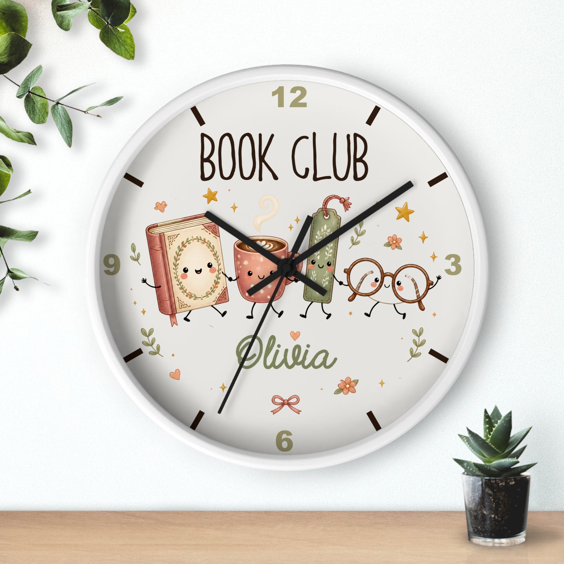 A round wall clock with a white frame and a whimsical book-themed face, mounted on a white wall above a light wood surface. The clock features the words 'BOOK CLUB' and the name 'Olivia' in a playful font, surrounded by cute illustrated books, glasses, and stars.