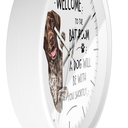 A white round wall clock with a printed design featuring a brown and white dog on the face, along with the text 'WELCOME TO THE BATHROOM A DOG WILL BE WITH YOU SHORTLY' and a paw print.