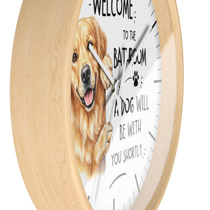 A round wall clock with a light natural wood frame and a white face, featuring a golden retriever illustration and the text 'WELCOME TO THE BATHROOM A DOG WILL BE WITH YOU SHORTLY'.