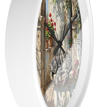 Round white wall clock featuring a printed illustration of a gray tabby cat lounging on a sunlit stone balcony with red flowers and green vines.