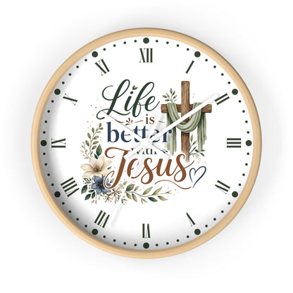 A round wall clock with a light wood frame and a white face, featuring the text 'Life is better with Jesus' in decorative script and serif fonts, surrounded by floral and leaf illustrations and a wooden cross graphic.