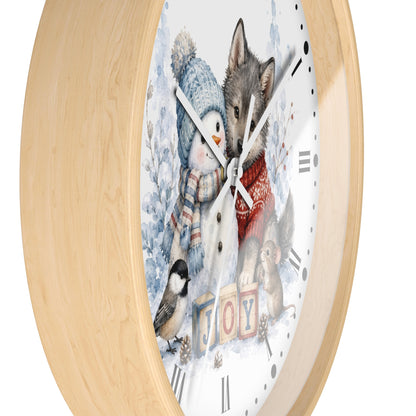 A round wall clock with a light natural wood frame and a white face featuring a winter illustration of a snowman, a wolf, a penguin, a chickadee, and the word "JOY" in colorful blocks.