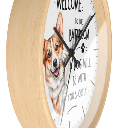 A round wall clock with a light natural wood frame and a white face. The clock face features a painted corgi illustration and the humorous text 'WELCOME TO THE BATROOM A DOG WILL BE WITH YOU SHORTLY'.
