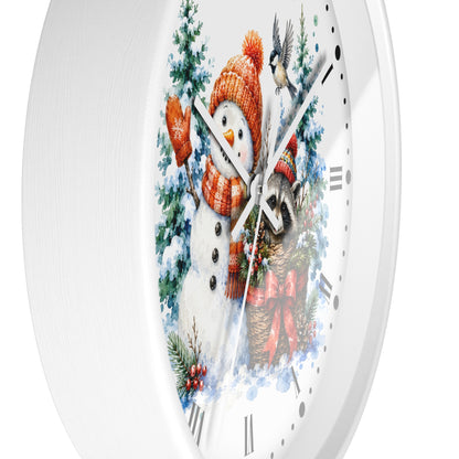 A white round wall clock with a festive winter illustration on the face, featuring a snowman in an orange knit hat and scarf, evergreen trees, a bird, and a raccoon near a red bow, shown against a white background.