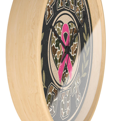 A close-up of a round wall clock with a light natural wood frame and a printed dial featuring a pink ribbon over a camouflage heart, set against a plain white background.