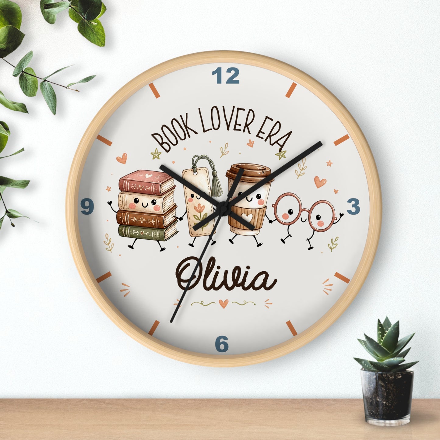 A round wall clock with a light wood frame and a white face, featuring cute cartoon illustrations of books, a coffee cup, and reading glasses. The clock is personalized with the text 'BOOK LOVER ERA' and the name 'Olivia' in black script.