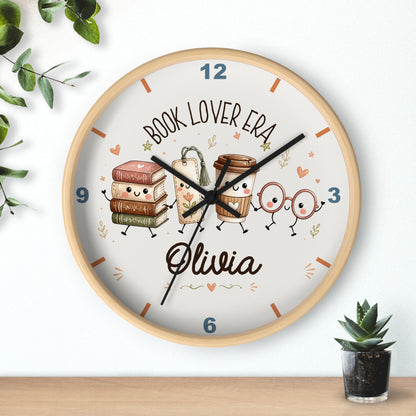 A round wall clock with a light wood frame and a white face, featuring cute cartoon illustrations of books, a coffee cup, and reading glasses. The clock is personalized with the text 'BOOK LOVER ERA' and the name 'Olivia' in black script.