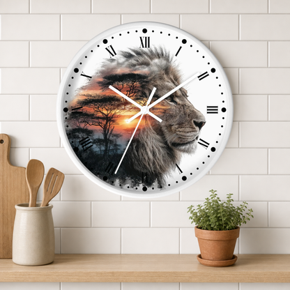 A round wall clock with a white face and black Roman numerals, featuring a printed design of a lion's face over a sunset savanna scene. The clock is mounted on a white subway tile wall above a wooden shelf with a small potted plant and kitchen utensils.