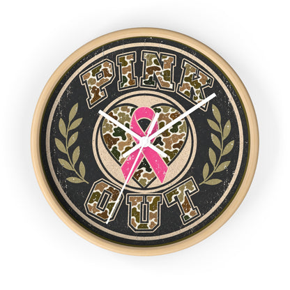 A round wall clock with a distressed design featuring the words 'PINK OUT' in camouflage lettering, a pink ribbon heart in the center, and olive branch motifs on a black background with a light wood-toned frame.