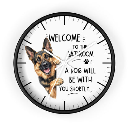 A round wall clock with a black frame and white face, featuring a German Shepherd illustration and the text 'WELCOME TO THE BATHROOM A DOG WILL BE WITH YOU SHORTLY.' on a light gray background.