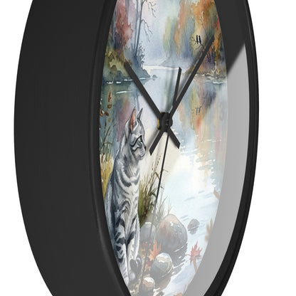 A round wall clock with a black frame and a printed watercolor-style face showing a gray tabby cat beside a reflective lake in autumn colors. The clock is photographed against a plain white background.