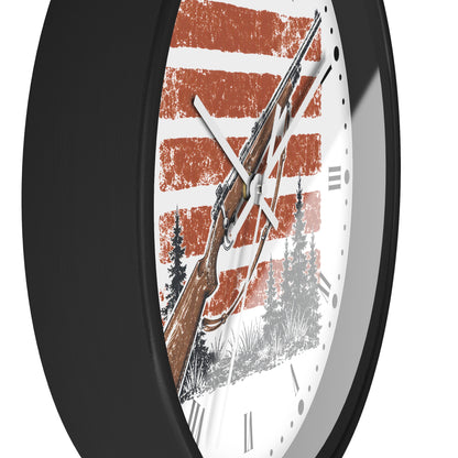 A close-up of a round wall clock with a black frame and a white face featuring a rustic orange and white striped design, a brown rifle illustration, and black tree silhouettes.