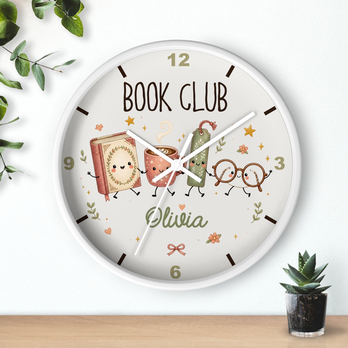 A round wall clock with a white frame and a whimsical 'Book Club' design featuring cartoon characters and the name 'Olivia'. The clock is shown hanging on a white wall above a wooden shelf with a small potted succulent and decorative leaves.