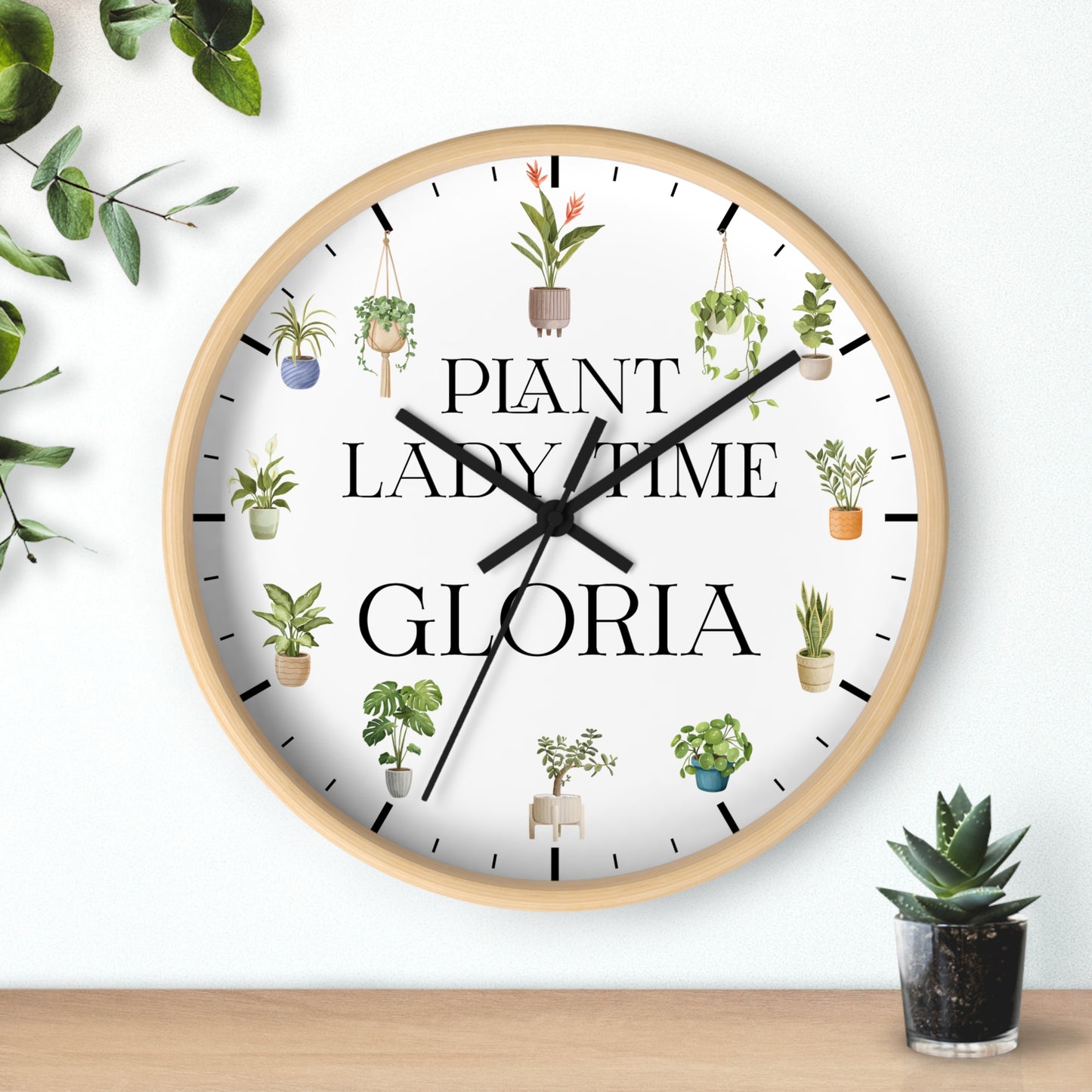 A round wall clock with a light wood frame and a white face, featuring the text 'PLANT LADY TIME GLORIA' in black serif font and small illustrated potted plants around the dial. The clock is shown hanging on a white wall above a wooden surface, with decorative greenery in the corners.