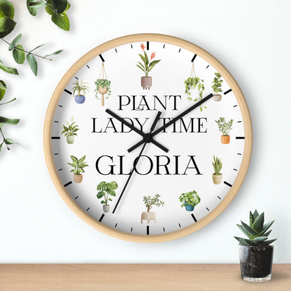 A round wall clock with a light wood frame and a white face, featuring the text 'PLANT LADY TIME GLORIA' in black serif font and small illustrated potted plants around the dial. The clock is shown hanging on a white wall above a wooden surface, with decorative greenery in the corners.