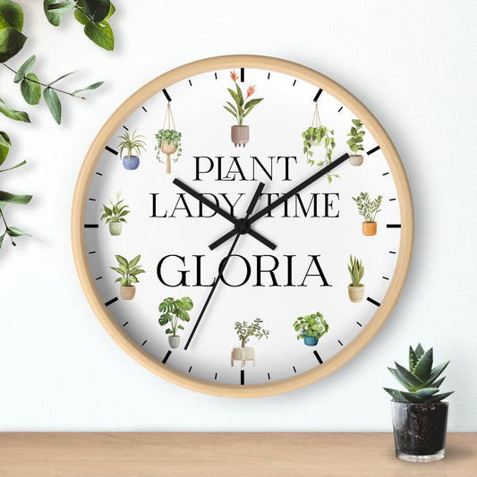 A round wall clock with a light wood frame and a white face, featuring the text 'PLANT LADY TIME GLORIA' in black serif font and small illustrated potted plants around the dial. The clock is shown hanging on a white wall above a wooden surface, with decorative greenery in the corners.
