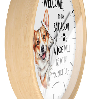 A round wall clock with a light natural wood frame and a white face. The clock face features a painted corgi illustration and the humorous text 'WELCOME TO THE BATROOM A DOG WILL BE WITH YOU SHORTLY'.