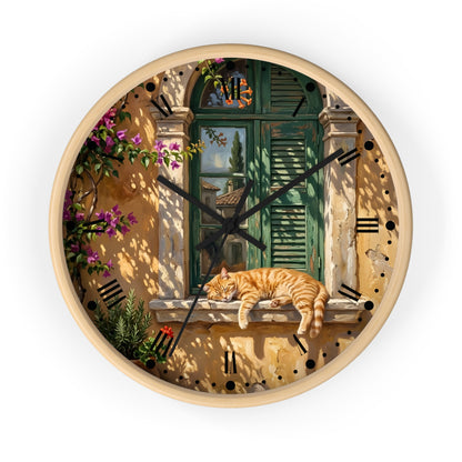 Round wall clock with a light wood frame and a printed scenic design featuring a green shuttered window, warm beige walls, black Roman numerals, and an orange tabby cat resting on a windowsill. Black clock hands are visible against a plain white background.