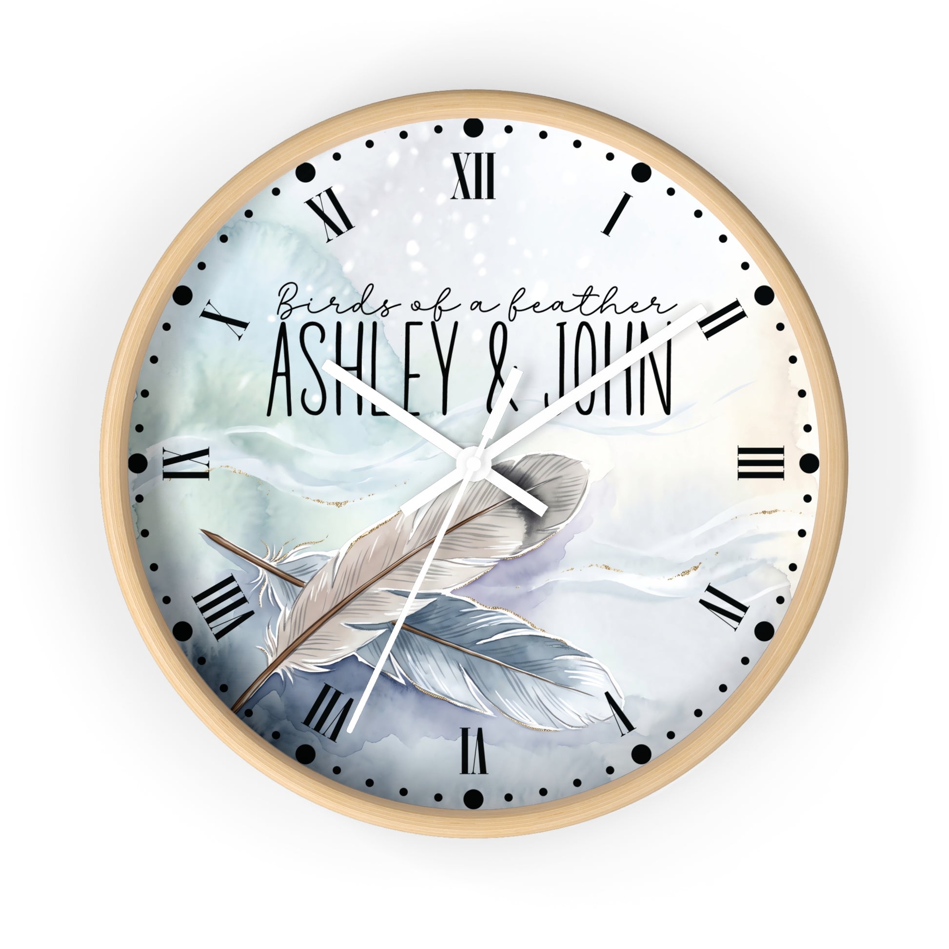 A round wall clock with a light wood frame and a custom printed dial featuring the text 'Birds of a feather ASHLEY & JOHN' over a watercolor illustration of two feathers in soft blue, white, and gray tones. The clock has black Roman numerals and white hands, shown against a plain white background.