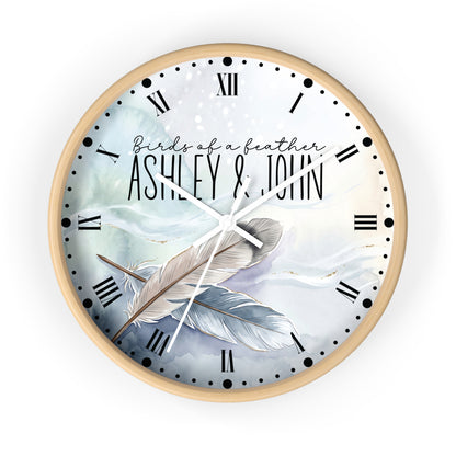 A round wall clock with a light wood frame and a custom printed dial featuring the text 'Birds of a feather ASHLEY & JOHN' over a watercolor illustration of two feathers in soft blue, white, and gray tones. The clock has black Roman numerals and white hands, shown against a plain white background.