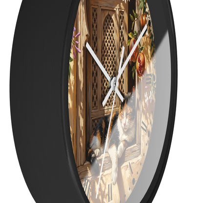 A round wall clock with a black frame and a printed scene on the face showing a calico cat resting by a wooden door, photographed against a plain white background.