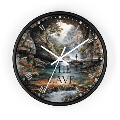 A round wall clock with a black frame and a scenic landscape print featuring a cave opening over a stream, trees, rocks, and the text 'THE CAVE'. White clock hands and Roman numeral hour markers are visible against a plain white background.