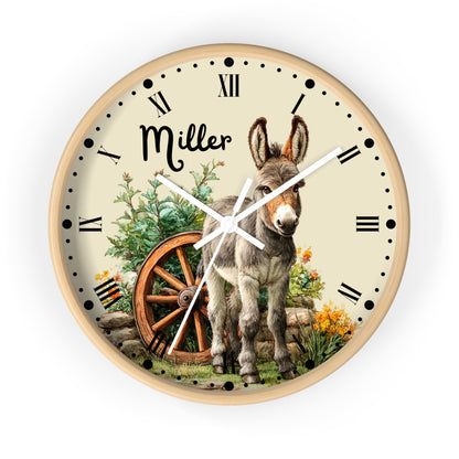 A round wall clock with a light wood frame and a cream dial, featuring a printed illustration of a donkey, a wagon wheel, flowers, and the name 'Miller' in script. The clock is shown against a plain white background.