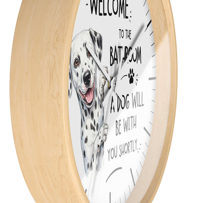 Round wall clock with a light natural wood frame and a white face featuring a Dalmatian illustration and the text 'WELCOME TO THE BATROOM A DOG WILL BE WITH YOU SHORTLY'.