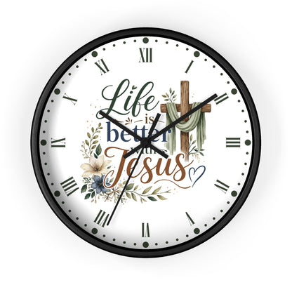 A round wall clock with a black frame and a white face featuring the text 'Life is better with Jesus' in decorative script, Roman numerals, floral illustrations, and a wooden cross.