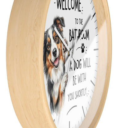 Round wall clock with a light natural wood frame and a white face featuring a watercolor illustration of a tricolor dog and the text 'WELCOME TO THE BATHROOM A DOG WILL BE WITH YOU SHORTLY'.