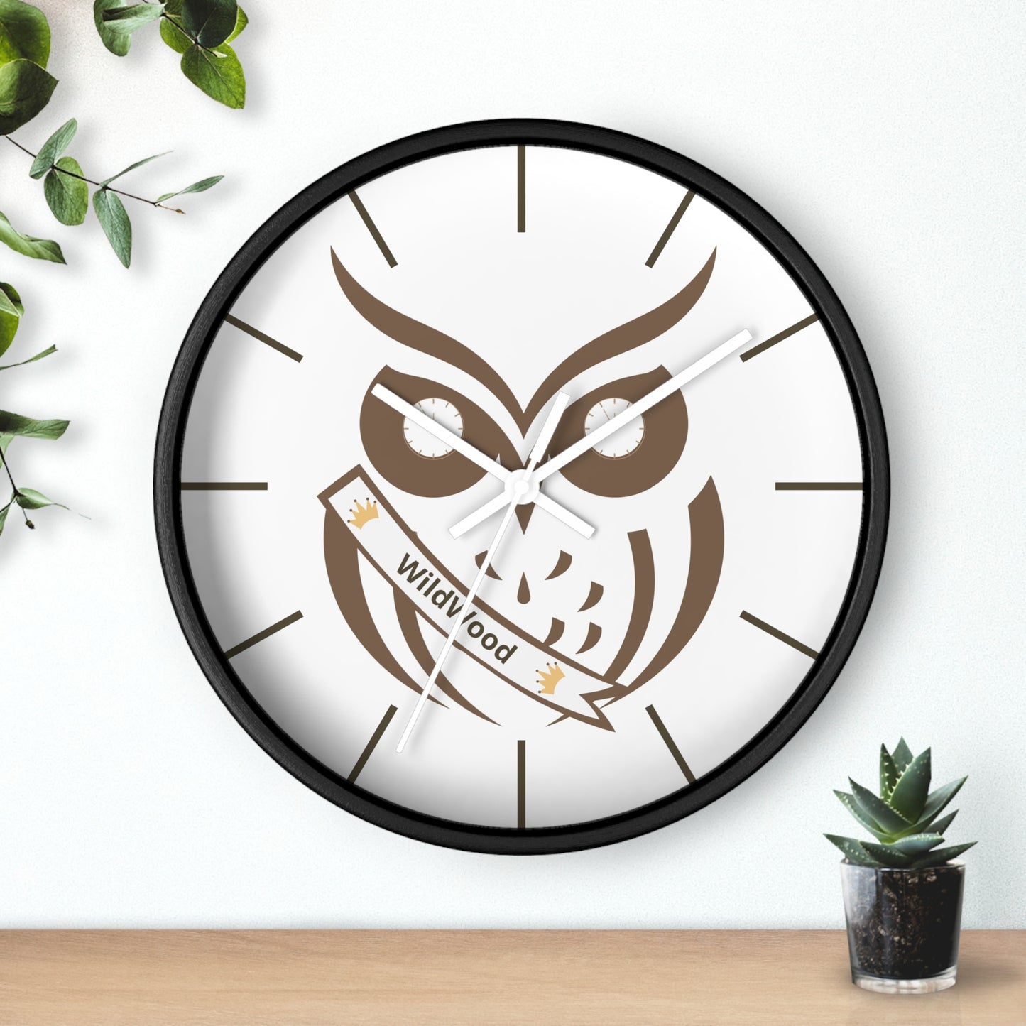 A round wall clock with a black frame and a white face featuring a brown owl illustration and a banner that reads 'Wild Good'. The clock is mounted on a white wall above a light wood surface, with green leaves and a small potted succulent as decorative props.