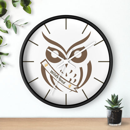 A round wall clock with a black frame and a white face featuring a brown owl illustration and a banner that reads 'Wild Good'. The clock is mounted on a white wall above a light wood surface, with green leaves and a small potted succulent as decorative props.