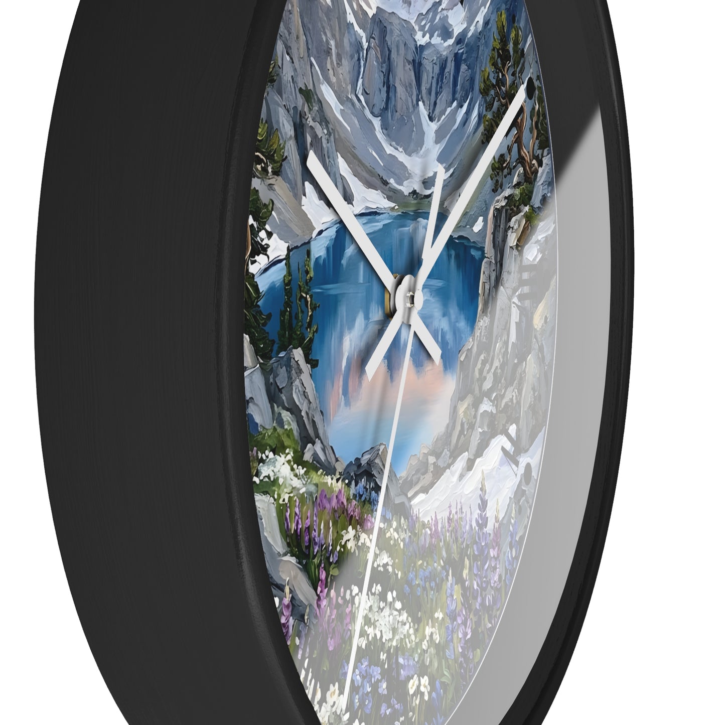 A round wall clock with a black frame and a scenic landscape print featuring a blue mountain lake, snowy peaks, trees, and wildflowers. The clock is shown at an angle against a plain white background.