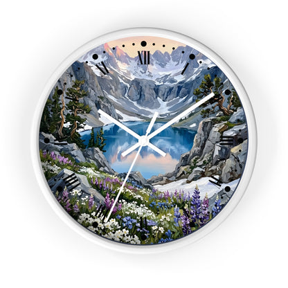 A round wall clock with a white frame and a scenic mountain landscape printed on the face. The artwork shows snowy peaks, a blue lake, rocky cliffs, evergreen trees, and wildflowers, with black Roman numerals and white clock hands.